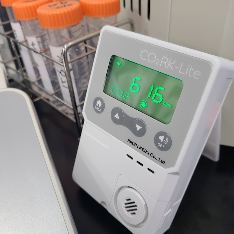 理研計器製のCO2モニター「CO2RK-Lite」が実験室の机に置かれている。緑色のデジタル画面にはCO2濃度616ppmと表示されている。背景には試験管立てに立てられたチューブが見える。